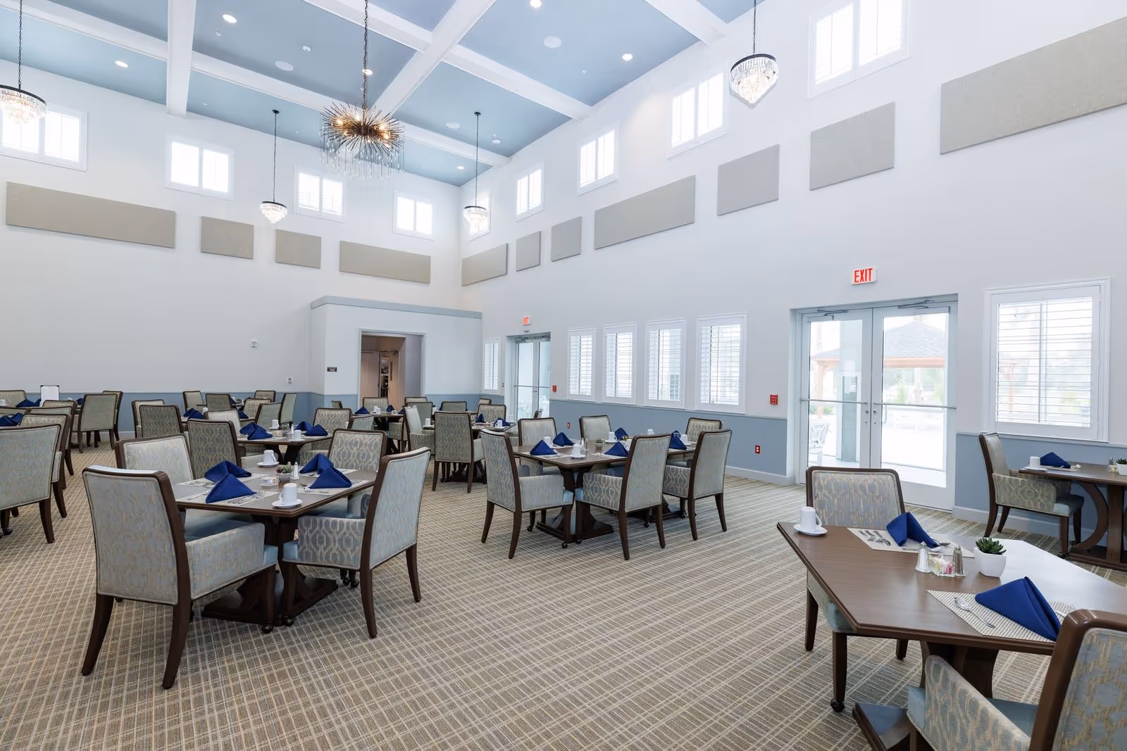 A spacious, well-lit dining room with multiple tables set with blue napkins, white cups, and small plants. The room features high ceilings with modern chandeliers, large windows allowing natural light, and a patterned carpeted floor.