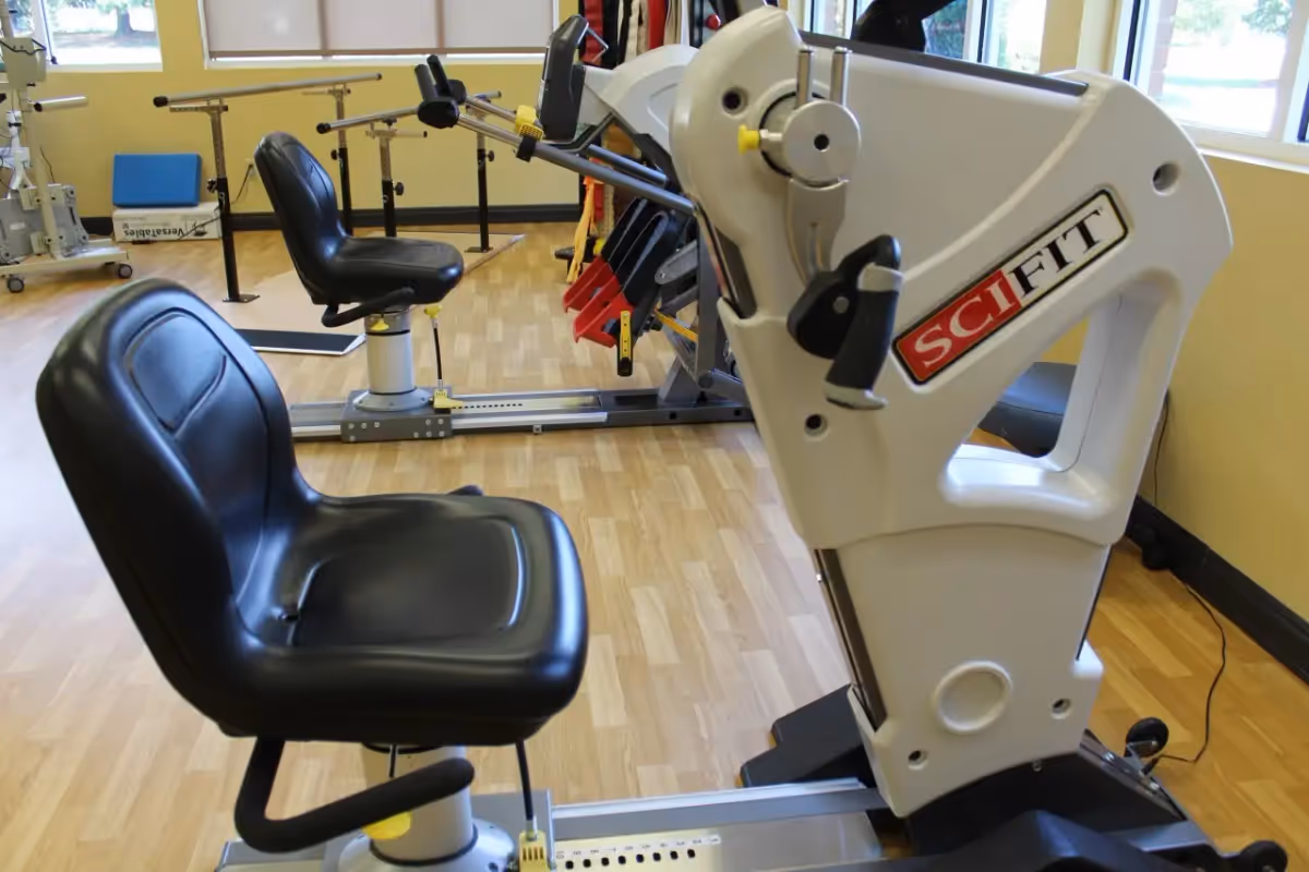 Exercise equipment with black padded seats and handles in a room with wooden flooring and yellow walls, featuring large windows letting in natural light.