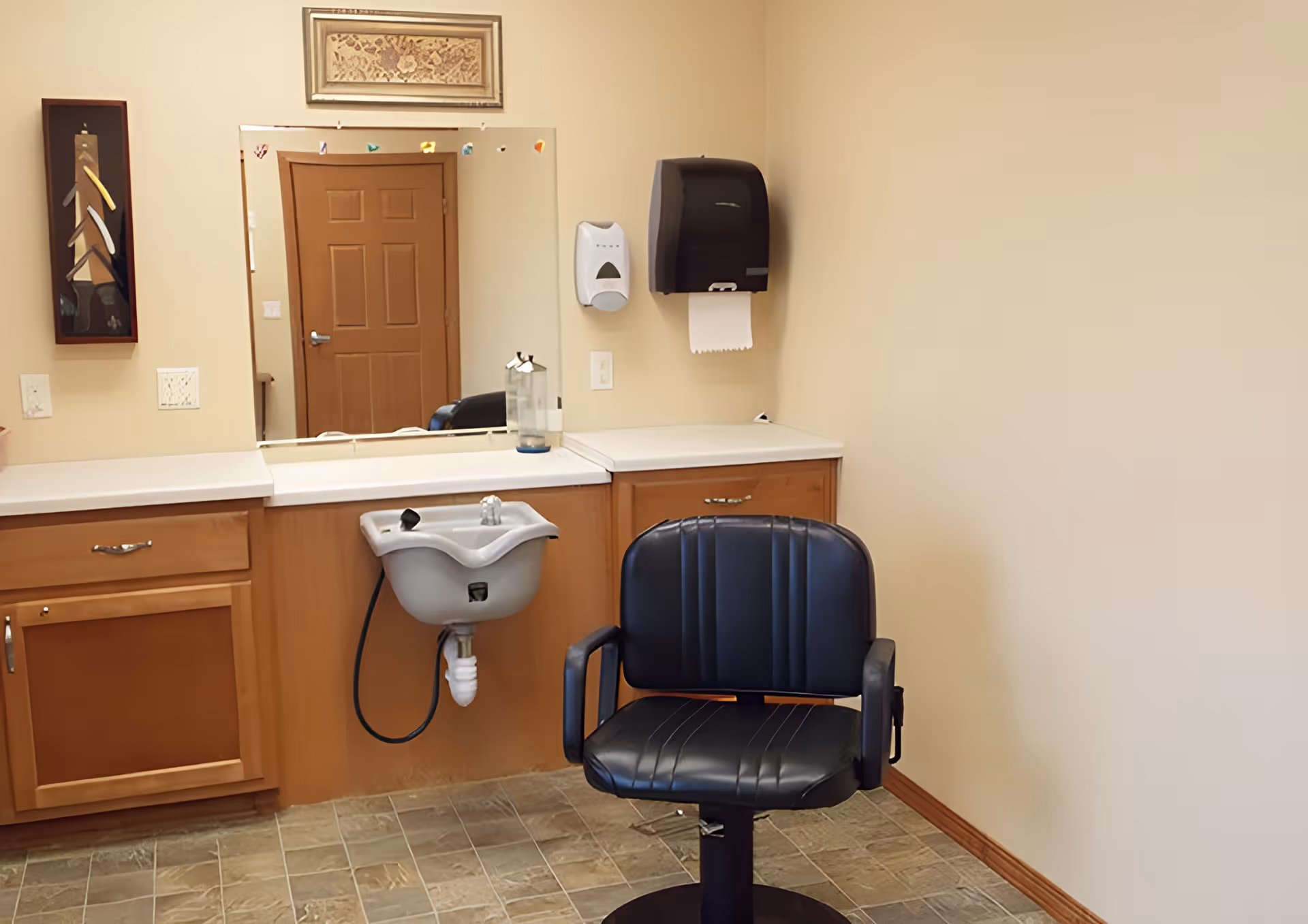 Interior salon station with a black barber chair in front of a small sink, countertop and mirror.