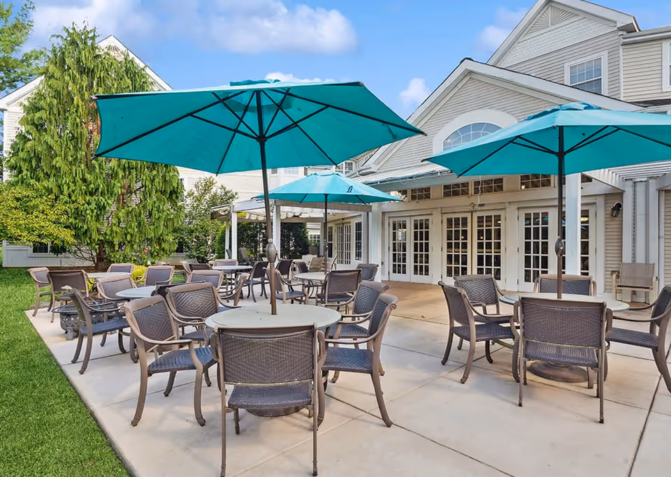 Outdoor patio area at Woodbridge Place Senior Living with multiple round tables, each surrounded by chairs and shaded by large turquoise umbrellas. The patio is adjacent to a building with large windows and doors, and there is green grass and trees nearby under a partly cloudy sky.