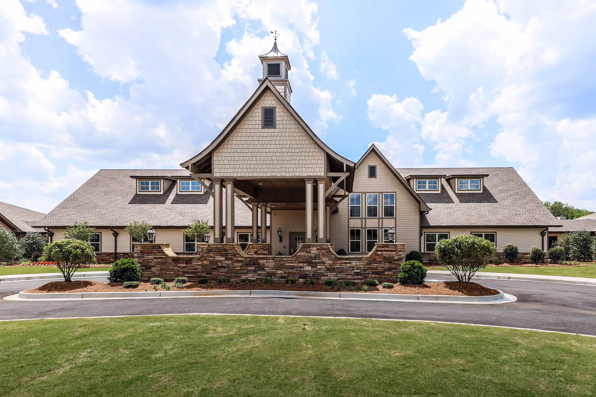 Front exterior view of a large senior living facility building with a covered entrance, stone accents, multiple windows, and a well-maintained lawn under a partly cloudy sky.
