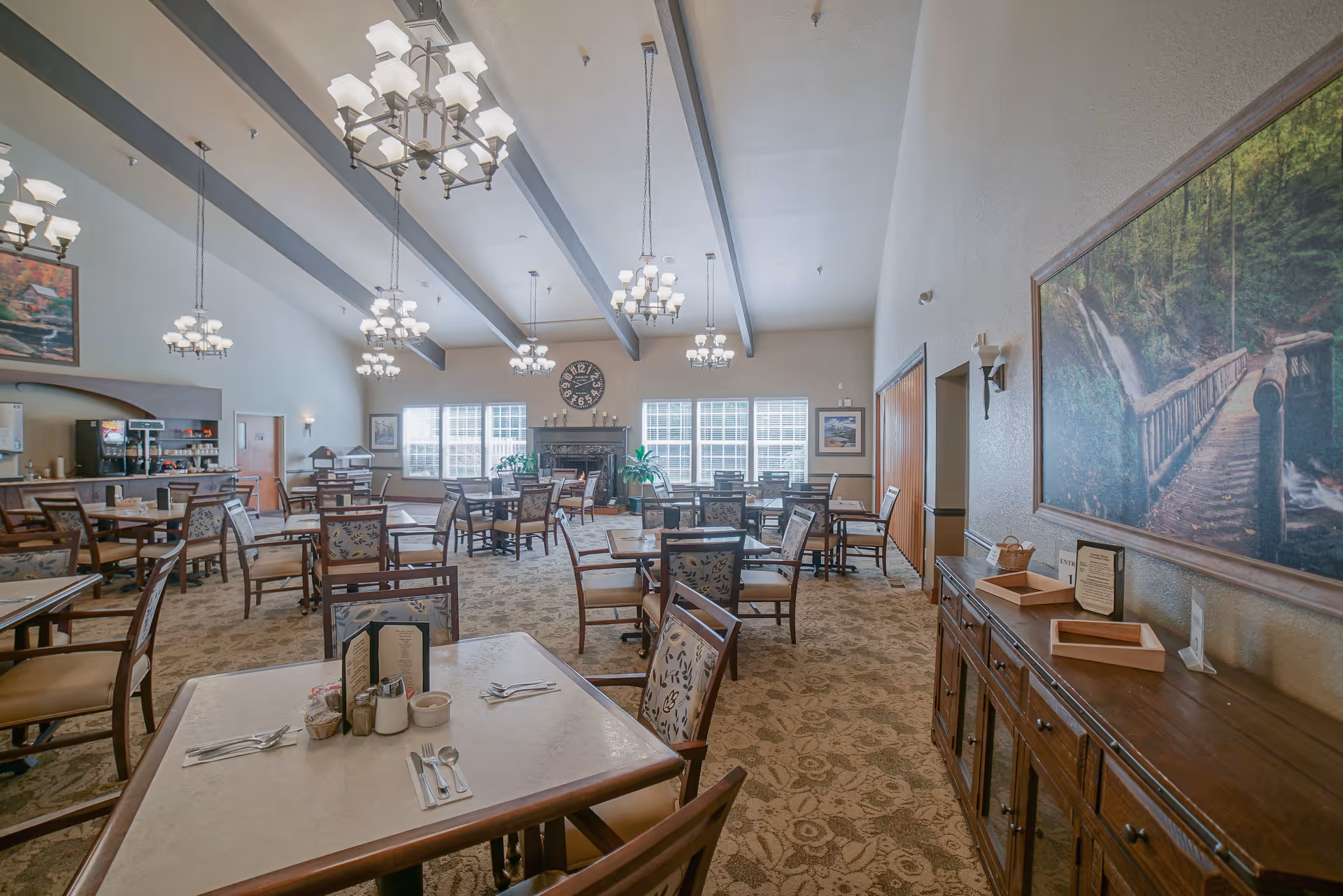 A spacious dining room with multiple tables and chairs arranged neatly. The room features high ceilings with exposed beams and several chandeliers providing warm lighting. There is a large clock above a fireplace at the far end, and large windows letting in natural light. On the right wall, there is a large framed picture of a forest scene with a wooden bridge. A wooden sideboard with trays and menus is also visible.