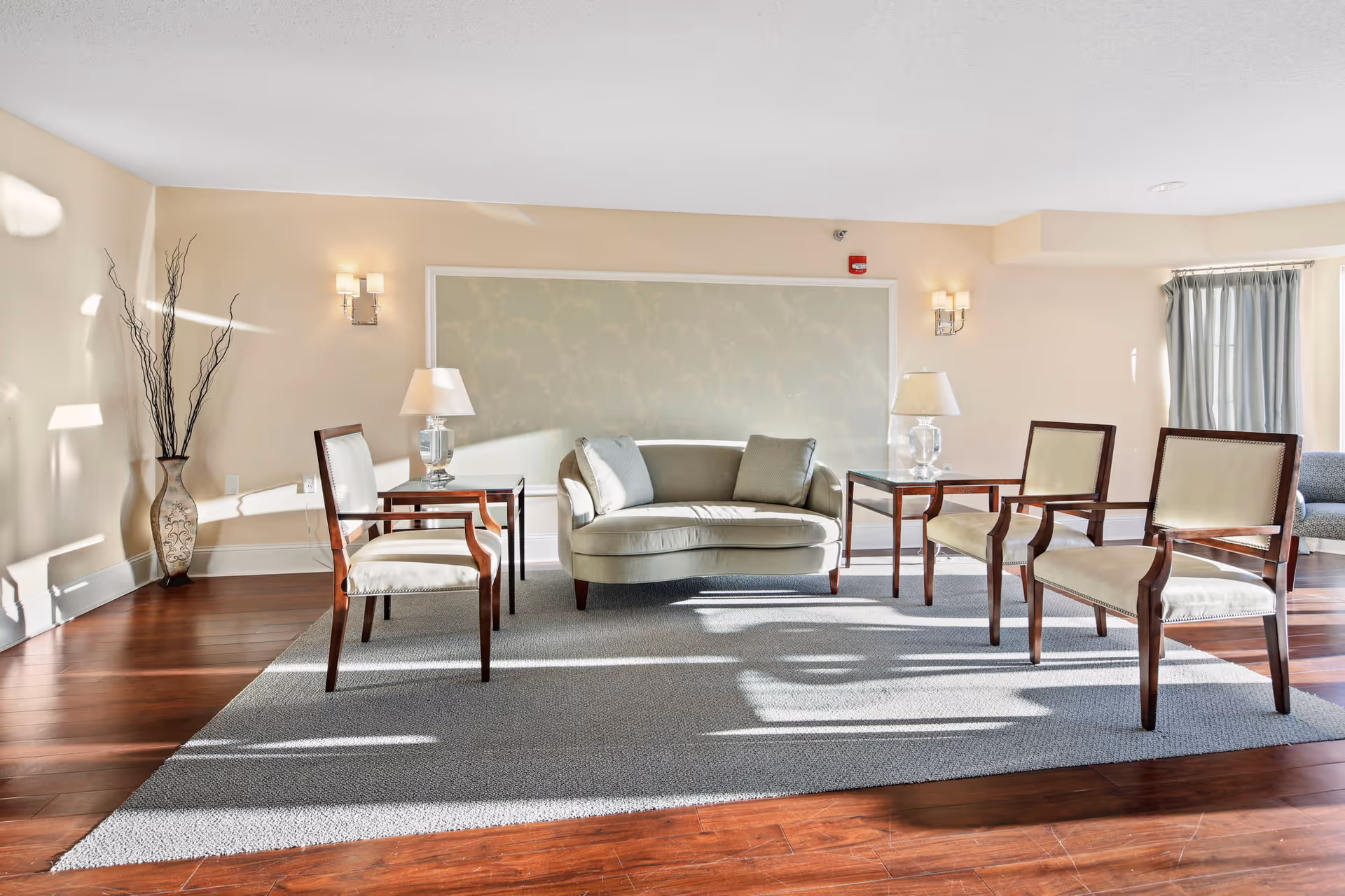 A bright and spacious living room area with wooden floors and a large gray rug. The room features a light green loveseat with two cushions, flanked by two glass table lamps on dark wooden side tables. There are four wooden chairs with cream-colored cushions arranged around the loveseat. A tall decorative vase with branches is placed in the corner, and natural light streams in through a window with gray curtains on the right side.