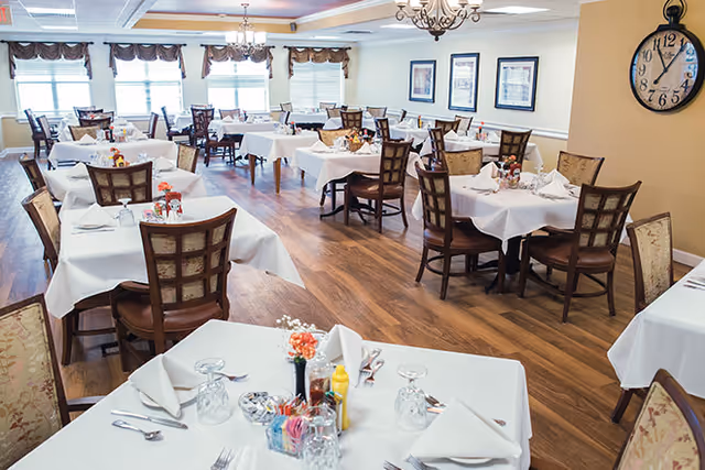 Bright formal dining room with multiple tables covered in white linens and set with glasses and silverware on a wood floor.