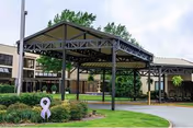 Covered entrance area of a senior living facility with a large metal canopy supported by black beams. The building in the background has multiple windows and beige walls. There is a landscaped garden with green bushes and a purple awareness ribbon decoration near the driveway.