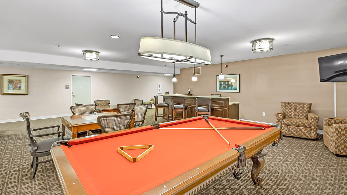 A game room featuring a red billiards table with a triangle rack, surrounded by chairs and tables. There is a bar area in the background with stools and a television on the wall.