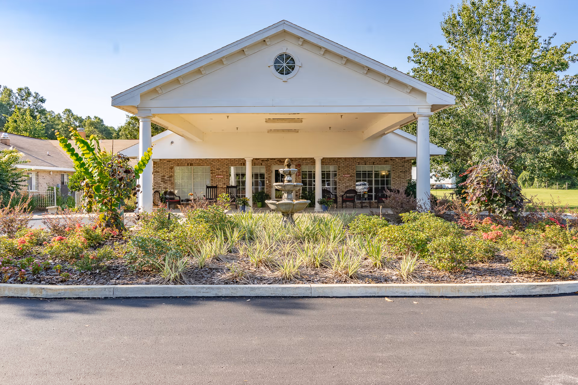 Front exterior view of Lafayette Nursing and Rehabilitation building with a covered entrance supported by white columns, a three-tiered water fountain in the landscaped garden area, and outdoor seating with chairs on the porch.
