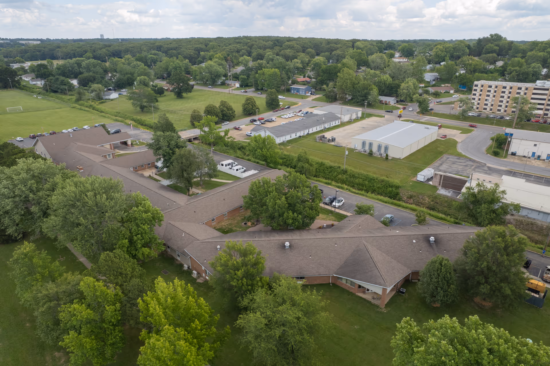Aerial view of a single-story senior living facility with connected wings surrounded by trees, lawns, parking areas and neighboring buildings.