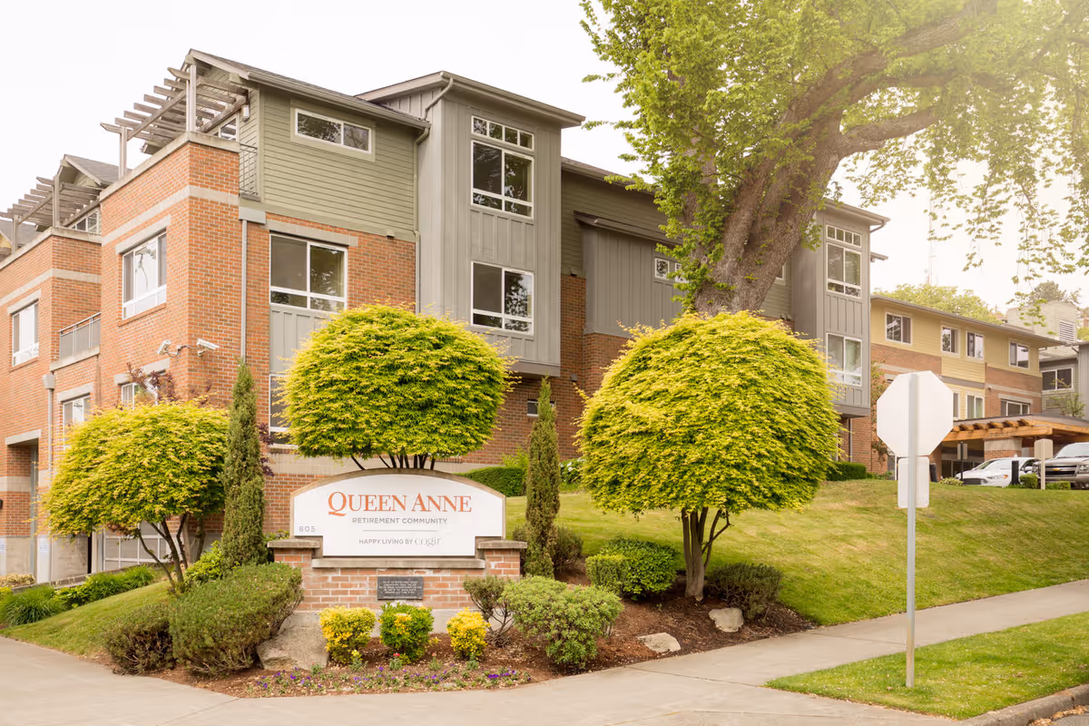 Exterior view of the Queen Anne Retirement Community building with well-maintained landscaping including trimmed bushes and trees. A sign in front reads 'Queen Anne Retirement Community, Happy Living by Cogir'. The building is a multi-story structure with brick and siding, and there is a sidewalk and street sign visible.