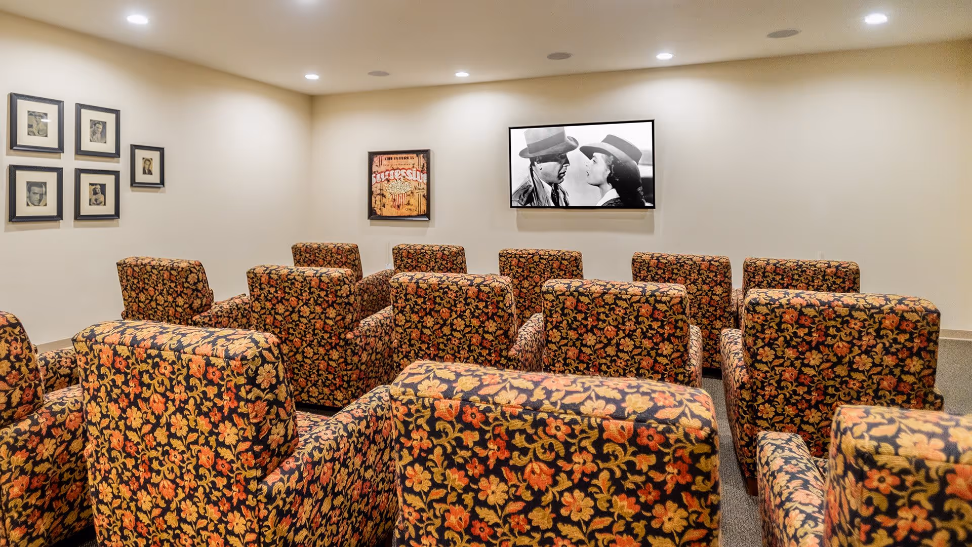 A small theater room with multiple rows of floral patterned armchairs facing a wall-mounted screen displaying a black and white movie scene. The room has beige walls adorned with framed pictures and posters.