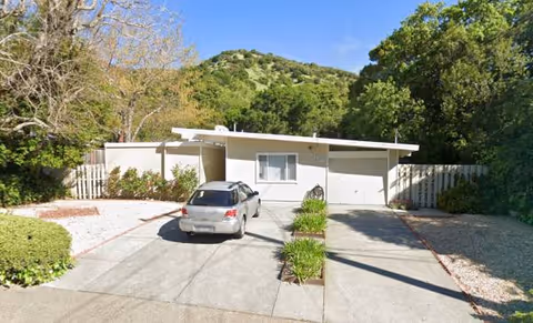 One-story residential-style building with a driveway, parked car, and green hills in background.