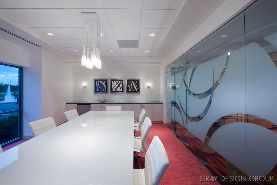 Modern conference room with a long white table surrounded by white chairs, a glass wall with frosted decorative patterns on the right, three abstract framed artworks on the back wall, and a window showing an outdoor fountain on the left.