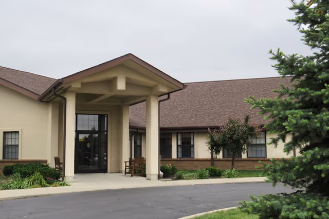 Exterior view of a single-story building with a covered entrance supported by two columns. The building has beige walls, brown roof shingles, and several windows. There is a paved driveway and some landscaping with bushes and a tree in the foreground.