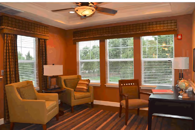 A cozy interior room with three chairs, two armchairs with cushions and one wooden chair, a striped carpet, a ceiling fan with light, two table lamps, large windows with blinds and curtains, and a desk with a globe and some books.