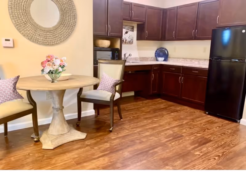 A kitchen area with dark wooden cabinets, a black refrigerator, and a countertop. In front of the kitchen is a round wooden table with a vase of flowers on top and two chairs with cushions. A decorative round mirror hangs on the wall above the table.