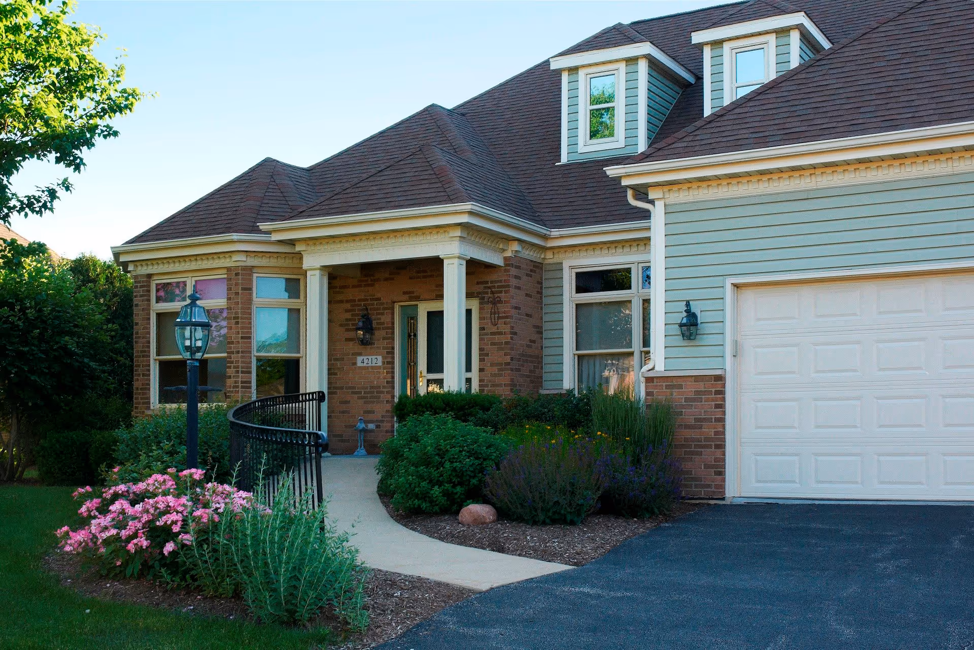 Front exterior view of a residential-style building with a brick and light blue siding facade, a dark shingled roof, a white garage door, a pathway leading to a covered entrance with white columns, surrounded by green bushes and pink flowers.