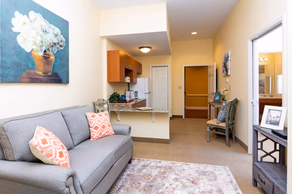 Interior view of a living area in a senior living facility with a gray sofa adorned with two patterned orange and white pillows, a floral painting on the wall above the sofa, a small kitchen area with wooden cabinets and a refrigerator in the background, a decorative chair and table with framed photos on the right, and a hallway leading to other rooms.