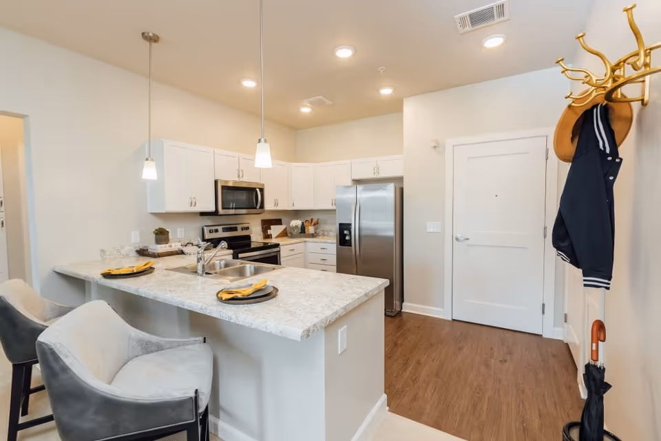 Modern kitchen with white cabinets, stainless steel refrigerator, stove, and microwave. A marble countertop island with two gray cushioned chairs and place settings with yellow napkins. A coat rack with a jacket and umbrella is visible near a white door. The floor is wood and the walls are light-colored.