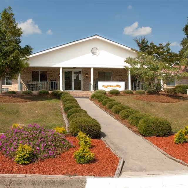Front exterior view of a single-story brick building with a white gabled roof and a covered porch. A concrete walkway lined with neatly trimmed bushes leads up to the entrance. The landscaping includes green shrubs, small trees, and a flower bed with purple and yellow flowers. The sky is clear with a few clouds.