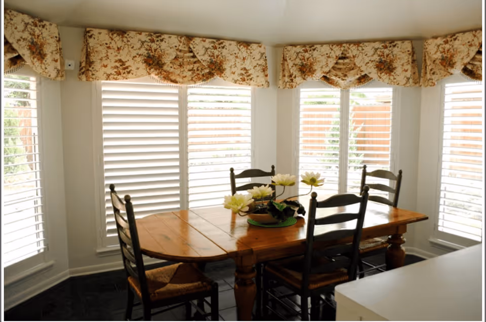 A dining area with a wooden table and four chairs. The table has a decorative centerpiece with white flowers. The room has large windows with white plantation shutters and floral valances.
