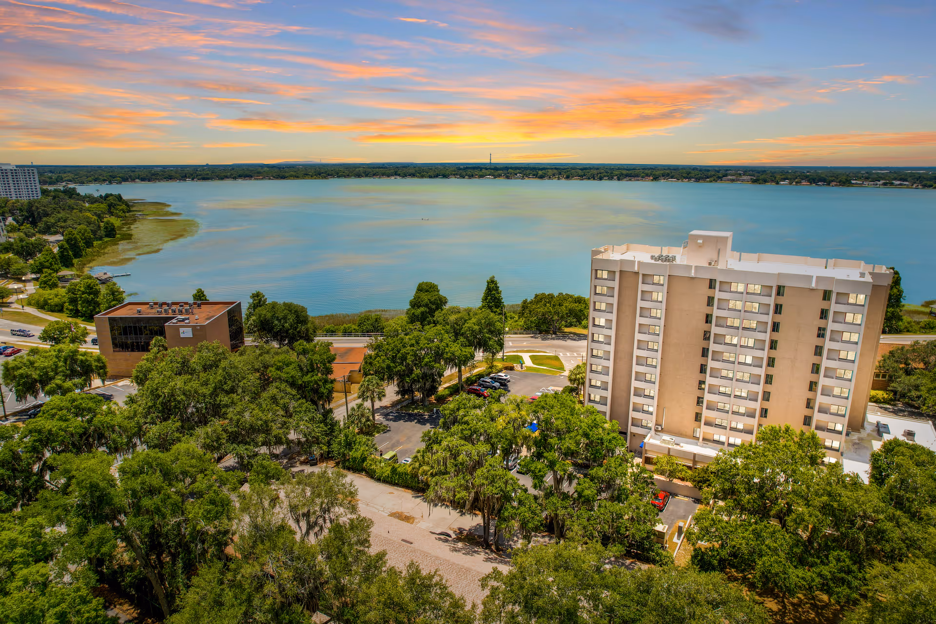 Aerial view of a multi-story residential building surrounded by trees near a large lake under a colorful sky at sunset.