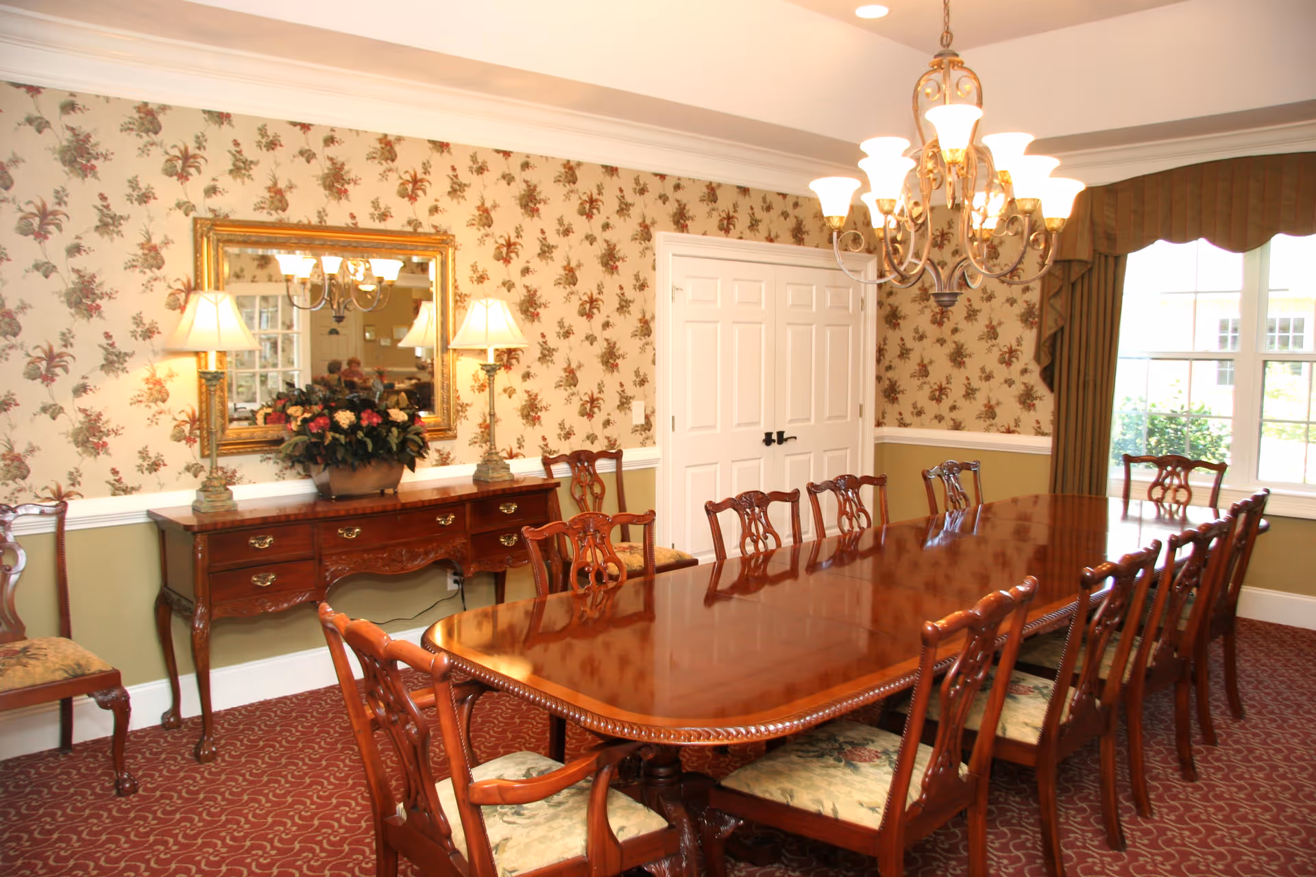 A formal dining room with a long polished wooden table surrounded by wooden chairs with floral upholstered seats. The walls have floral wallpaper above a white chair rail and green paint below. A large mirror with a gold frame hangs above a wooden sideboard with two lamps and a floral arrangement. A chandelier with multiple lights hangs above the table. There is a window with brown curtains letting in natural light.