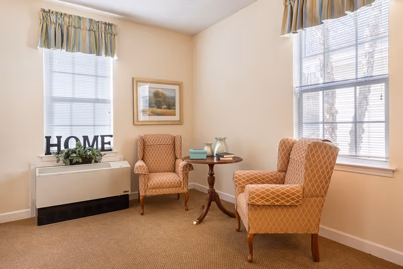 A cozy sitting area with two upholstered armchairs and a small round table between two windows.