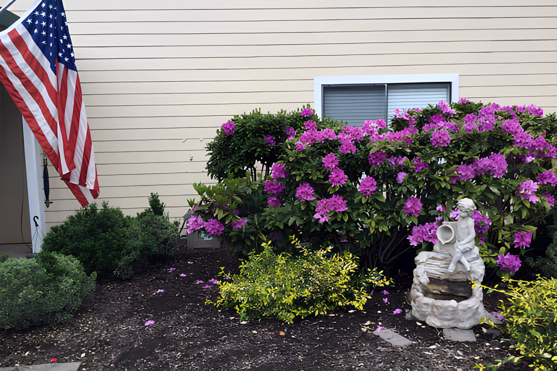 Outdoor garden area with green shrubs and vibrant purple flowers in front of a beige building wall with a window. An American flag is displayed on the left side near the entrance. A small white statue of a child holding a jug is placed among the plants.