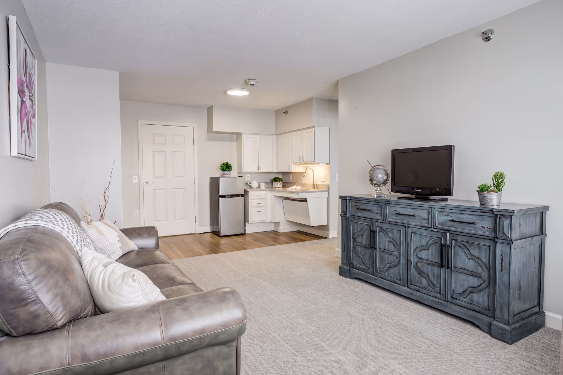 Interior view of a senior living facility unit featuring a gray leather sofa with pillows on the left, a small kitchenette with white cabinets, a mini refrigerator, and a sink in the background, and a blue distressed wooden cabinet with a flat-screen TV and decorative items on the right. The room has light gray walls and carpeted flooring with a wooden section near the kitchenette.