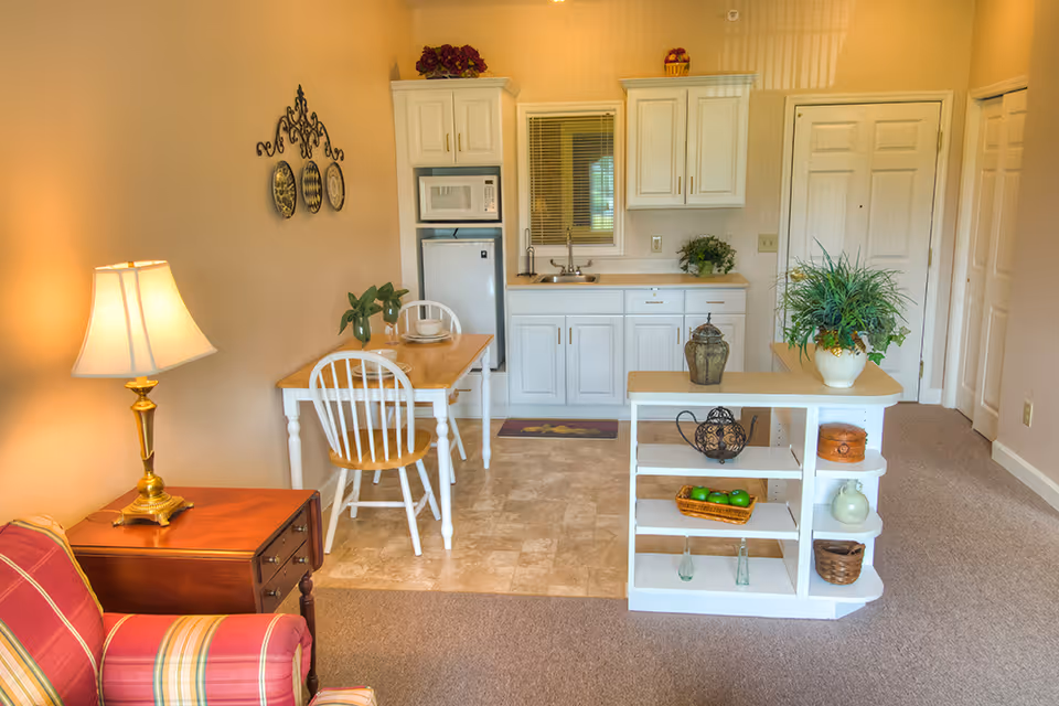 Open studio living area with a small kitchenette, dining table, shelving with decorative items, and a lamp-lit seating corner.