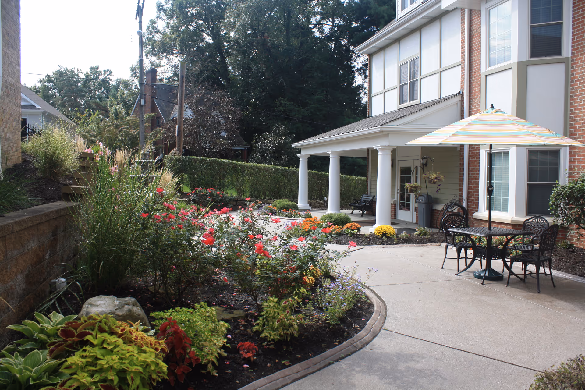 Outdoor patio area at New Hope Gracious Personal Care featuring a curved concrete walkway, colorful flower beds with various plants and flowers, a table with four chairs under a striped umbrella, and a building with white columns and large windows in the background.