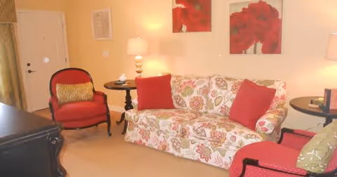 A bright living room with a floral-patterned sofa, red accent pillows and chairs, side tables with lamps, and red floral wall art.