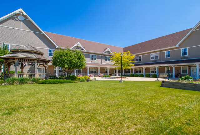 Exterior view of a senior living facility with a large green lawn, a wooden gazebo on the left, a tree with yellow leaves in the center, and a two-story beige building with a brown roof surrounding the courtyard under a clear blue sky.