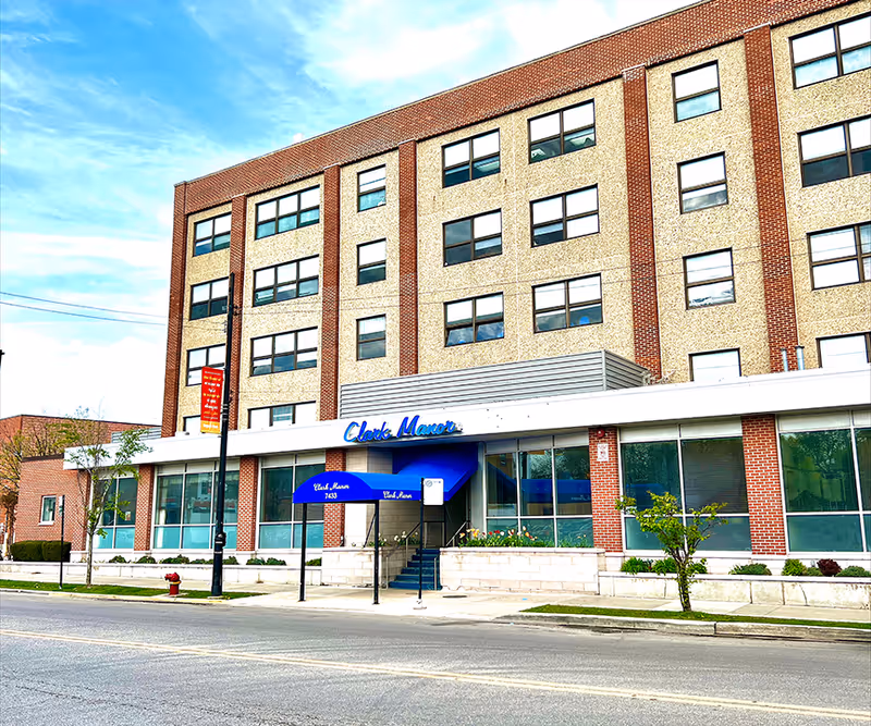 Exterior front view of Clark Manor, a multi-story brick and stone building with large windows and a blue awning over the entrance. The building is situated along a street with a sidewalk, small trees, and a fire hydrant visible.