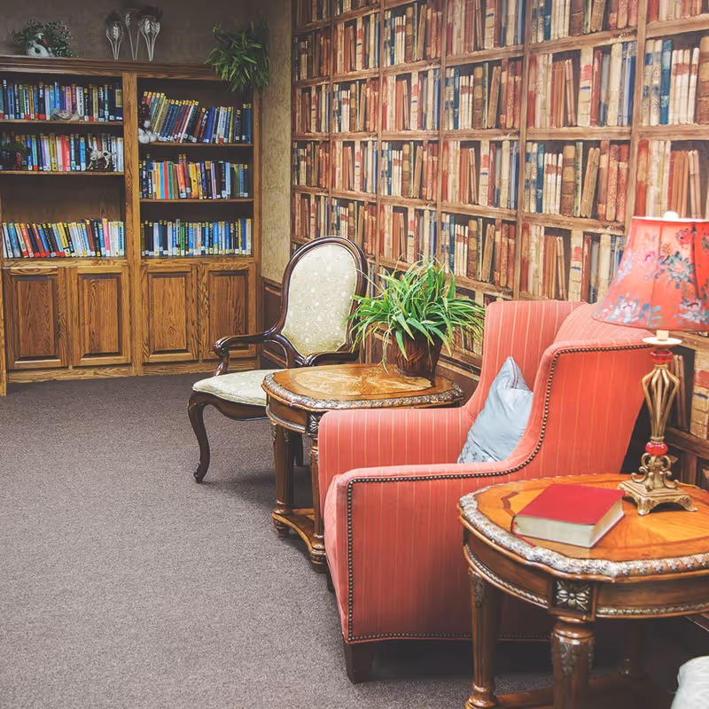 Cozy reading area in a senior living facility featuring a red upholstered armchair with a blue pillow, a wooden side table with a red book and a decorative lamp, a wooden chair with a cushioned seat, a small round wooden table with a potted plant, and bookshelves filled with books. The wall has wallpaper designed to look like bookshelves filled with old books.
