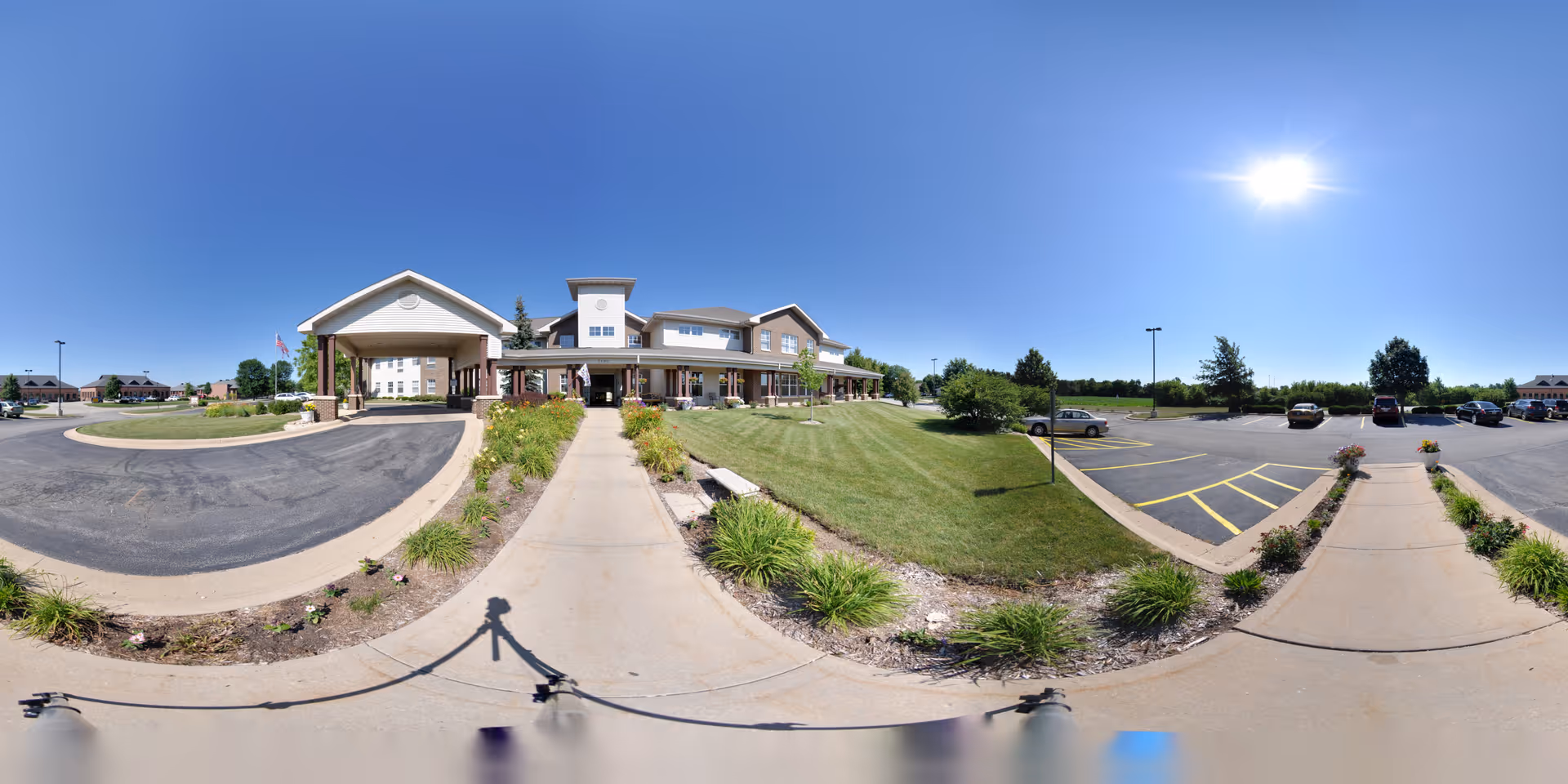 Wide panoramic view of the front exterior of Charter Senior Living of Rockford facility on a sunny day. The building has a covered entrance with columns, surrounded by landscaped greenery and a well-maintained lawn. There is a parking lot with several cars on the right side and a clear blue sky with the sun shining brightly.