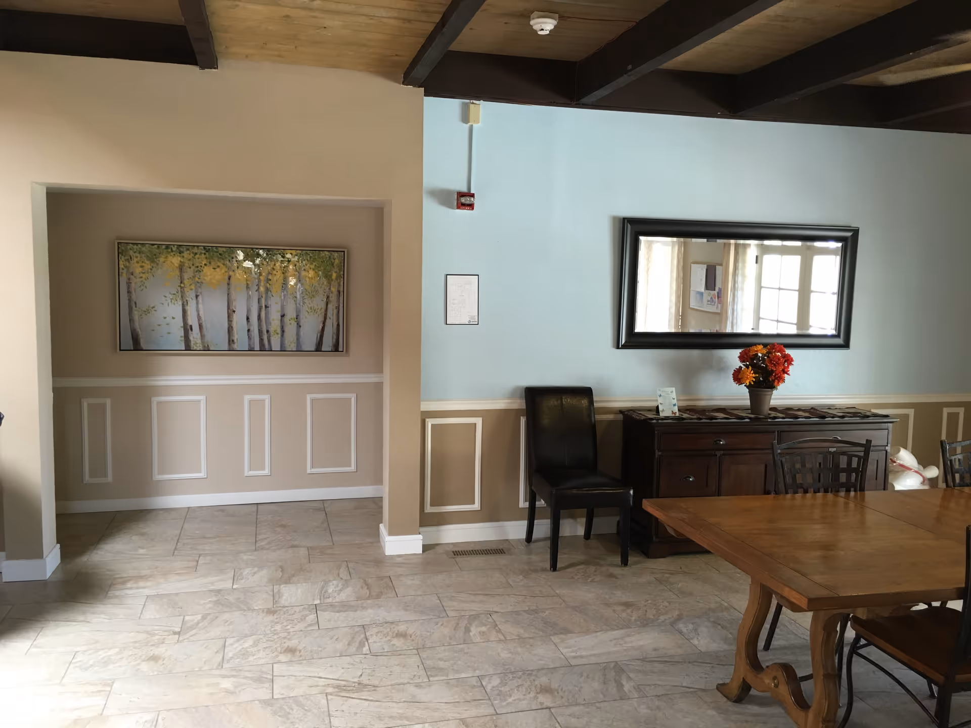 Interior view of a room with beige and light blue walls, a wooden ceiling with exposed beams, and tiled floor. There is a wooden dining table with chairs on the right, a dark wooden sideboard with a vase of flowers and a large rectangular mirror above it, and a black chair next to the sideboard. On the left, there is a recessed wall area with a framed painting of trees.