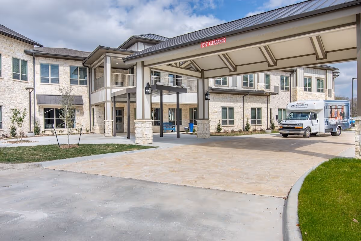 Entrance area of StoneCreek of Copperfield Senior Living facility showing a covered drop-off zone with a clearance sign, a two-story building with stone and siding exterior, windows, and a parked shuttle bus on the right side.