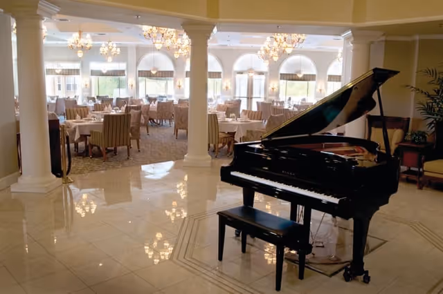 Elegant dining area with multiple tables and chairs arranged for guests, featuring large windows with arched tops allowing natural light to fill the room. In the foreground, there is a black grand piano with a matching bench on a polished tile floor. The space is decorated with chandeliers hanging from the ceiling and white columns separating the dining area from the piano space.