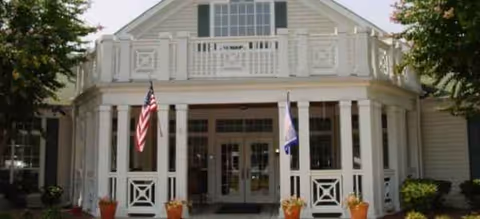 Front view of a light-colored senior living building with a covered porch and flags.