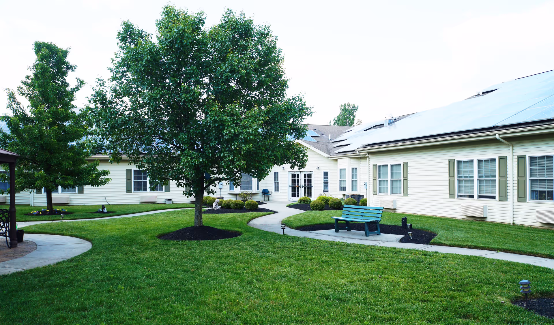 A well-maintained outdoor courtyard area at Traditions of Cross Keys featuring green grass, a few trees, a curved concrete pathway, a green bench, and a light yellow building with multiple windows in the background.