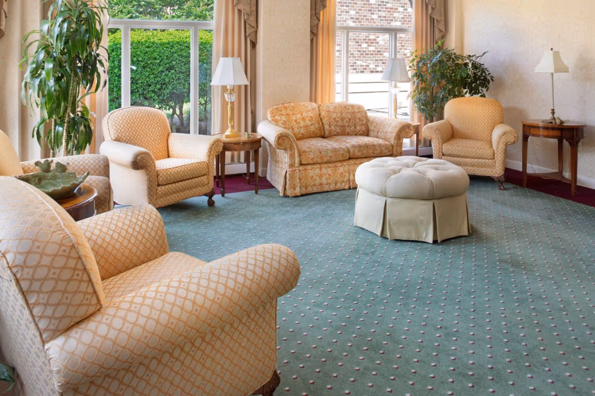 A cozy living room with patterned armchairs and a floral sofa arranged around a large, round, tufted ottoman. The room has green carpet with small red and white dots, two side tables with lamps, large windows with beige curtains, and green plants near the windows.
