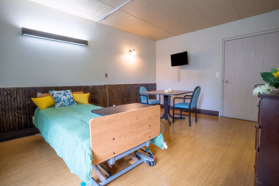 A nursing facility patient room with a single hospital bed, small table and chairs, wall-mounted TV, and a dresser with flowers.
