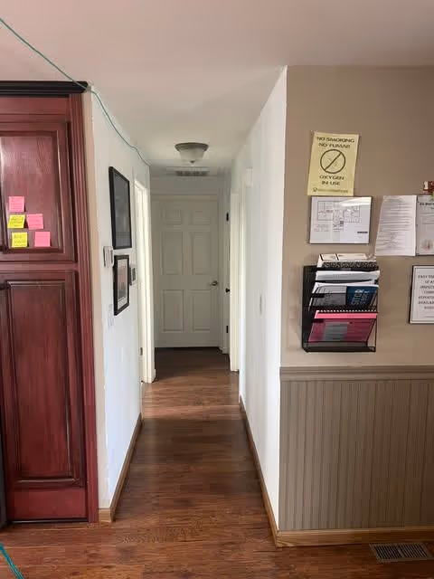 Narrow interior hallway with wood floors leading to a closed white door, red wooden cabinets on the left and bulletin rack with signs on the right wall.