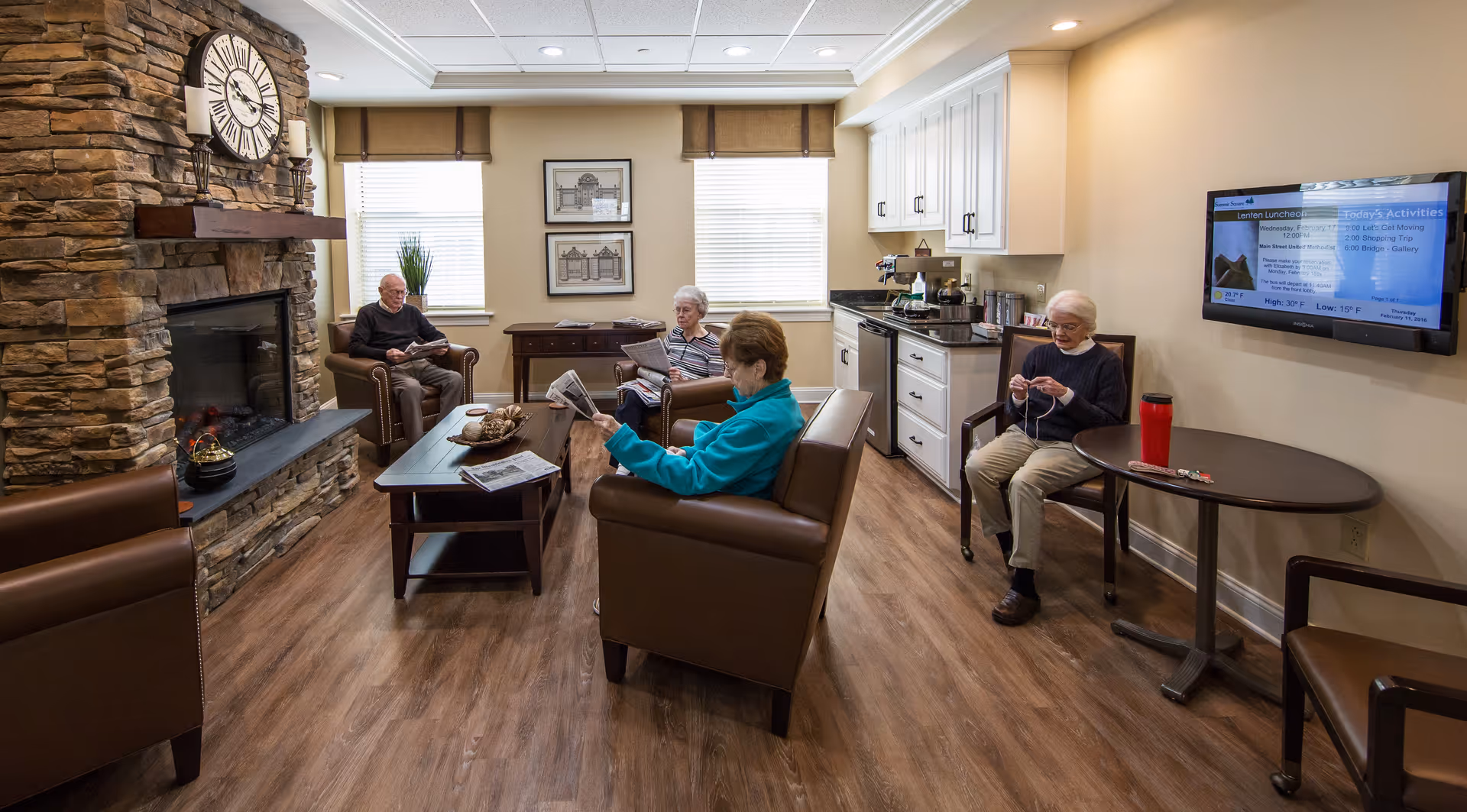 A cozy common area in a senior living facility with four elderly people engaged in different activities. Three are seated in brown leather armchairs near a stone fireplace with a large clock above it, reading newspapers. Another person is sitting at a round table knitting. The room has wood flooring, white cabinetry with a coffee station, two windows with blinds, framed pictures on the wall, and a flat-screen TV displaying activities and weather information.