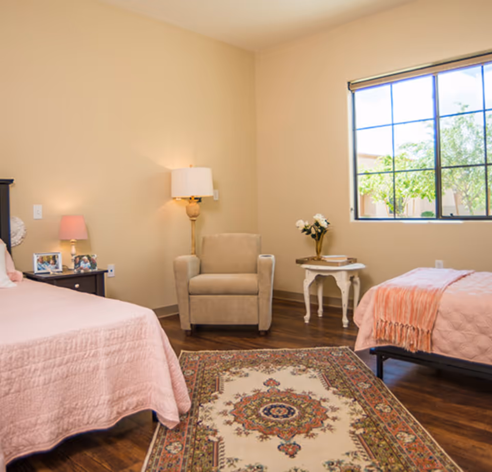 Two twin beds with pink quilts flank a seating area under a window in a bedroom.