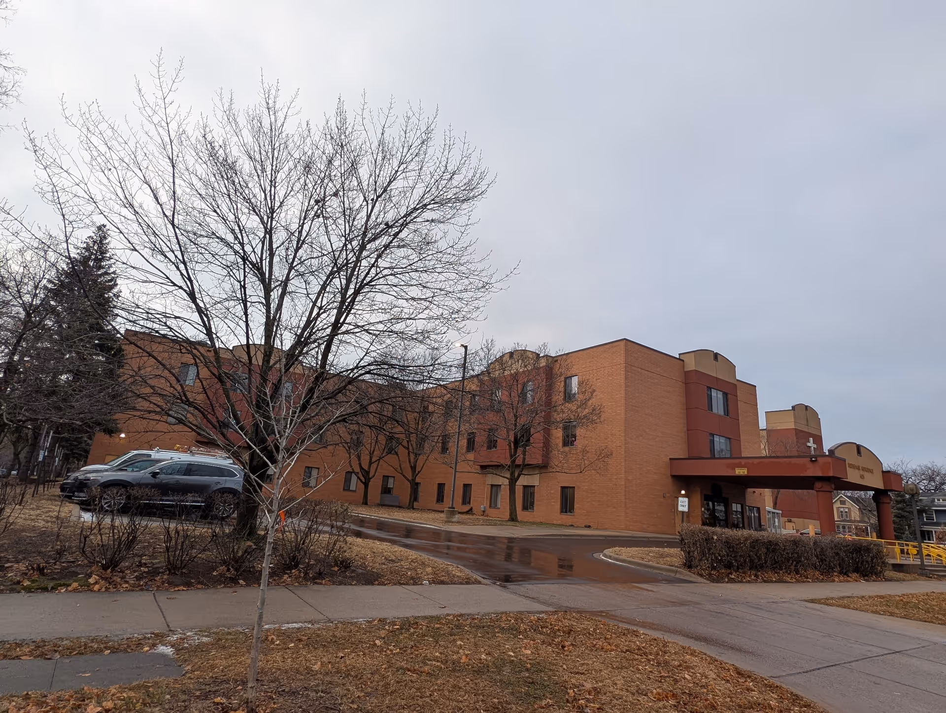 Brick three-story senior living facility with a covered entrance, parked cars, leafless trees, and an overcast sky.