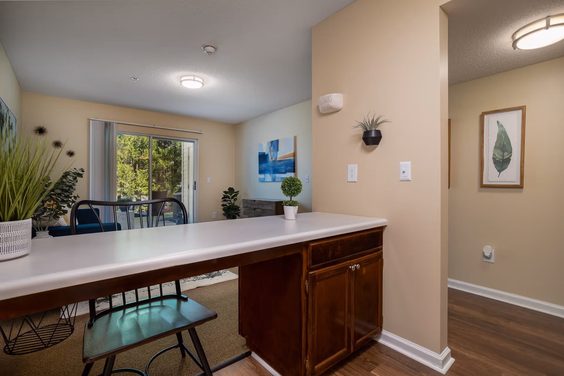Interior view of a living space in an assisted living facility showing a countertop with a wooden base and a chair in front. The room has beige walls, a sliding glass door leading outside, and decorative plants and artwork on the walls. The floor is a combination of carpet and wood.