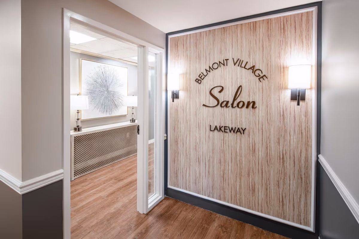 Interior hallway of Belmont Village Senior Living Lakeway showing an entrance to the salon. The wall features a wood-textured panel with the words 'Belmont Village Salon Lakeway' mounted on it, flanked by two wall sconces. The hallway has wood flooring and neutral-colored walls with white trim. Through the open door, a room with a decorative piece of art and two table lamps is visible.