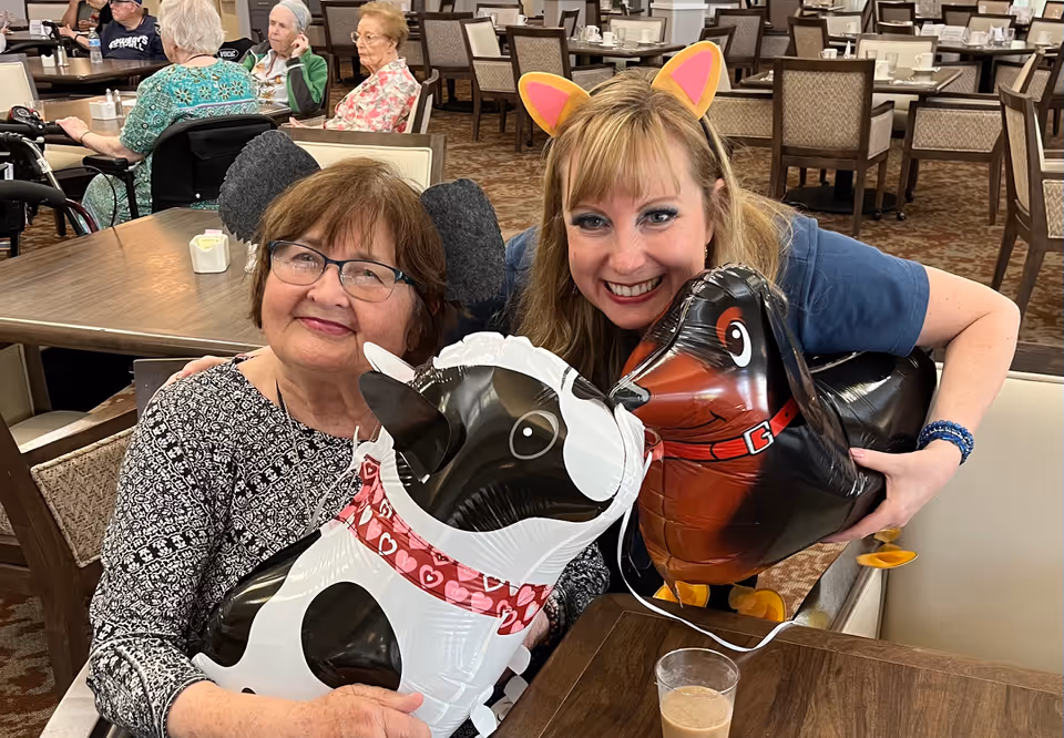 Two smiling women sit at a dining table in a senior living dining room holding dog-shaped balloons, with other residents and tables visible behind them.